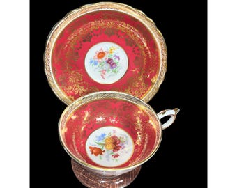 May include: A red teacup and saucer set with gold trim and floral designs. The cup and saucer are both decorated with a delicate floral pattern in the center, surrounded by a gold border and intricate gold designs.
