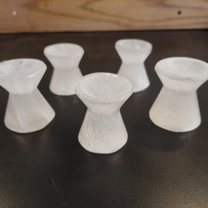 May include: Six white, hourglass-shaped candle holders. The holders have a wide, flat top and a flared base, with a narrower waist. The candle holders are made of a translucent material, possibly stone or glass.