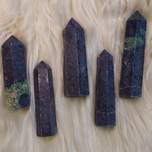 Puede incluir: Cinco puntas de piedras preciosas de color púrpura oscuro y verde, también conocidas como cristal o varita, están dispuestas sobre una superficie blanca y esponjosa.