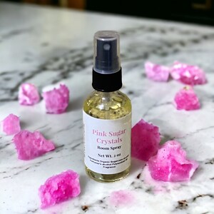 May include: A clear glass bottle with a black spray top filled with a light yellow liquid. The bottle has a white label with the text "Pink Sugar Crystals Room Spray Net Wt. 2 oz. Ingredients: Organic Sugarcane,  Perfumer's Alcohol, Polyethylene Glycol, Fragrance". The bottle is surrounded by pink sugar crystals.