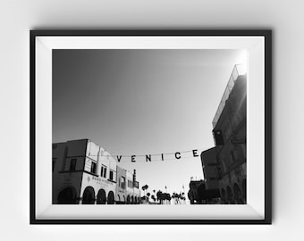 May include: Black and white photograph of a street scene in Venice, California. The word "VENICE" is spelled out in large black letters strung across the street. Buildings and palm trees are visible under a bright sky.