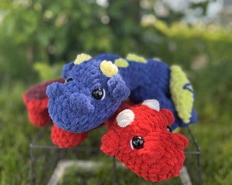 Puede incluir: Dos juguetes de dinosaurio de crochet, uno azul con detalles amarillos y otro rojo con manchas blancas. Los juguetes están sentados en un marco de alambre blanco frente a un fondo de hierba verde.