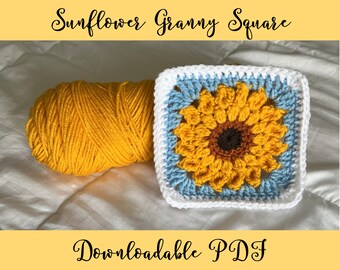 Può includere: Un quadrato di granny crochet a forma di girasole con un bordo blu e bianco. Il quadrato è realizzato con filato giallo, marrone e bianco. La matassa di filato è gialla.