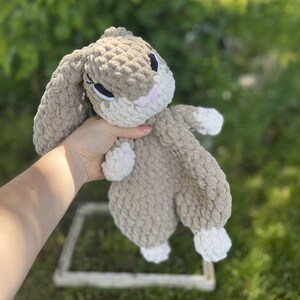 May include: A handmade, plush bunny in shades of tan and white. The bunny has long, floppy ears, black eyes, and a pink nose. The soft, textured material gives it a cuddly appearance, perfect for a child's toy or nursery decor.