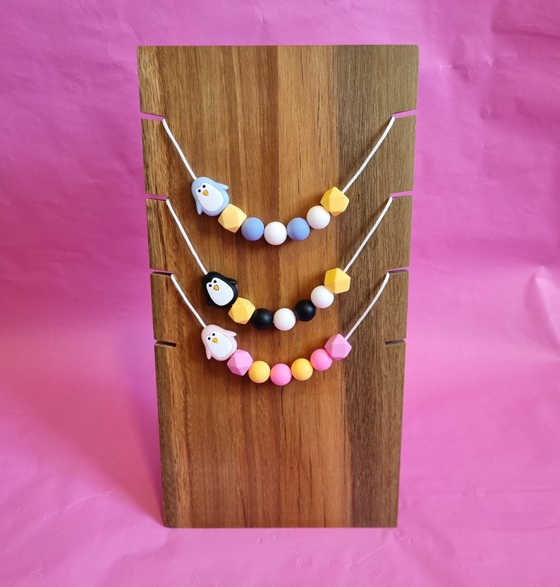 Puede incluir: Tres collares de silicona con dijes de ping&uuml;ino. Los collares se muestran en un soporte de madera. El collar superior tiene cuentas de color azul claro, gris y amarillo. El collar del medio tiene cuentas de color negro, blanco y amarillo. El collar inferior tiene cuentas de color rosa, amarillo y rosa claro.