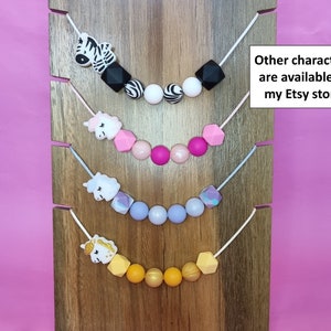 Puede incluir: Cuatro collares de dentici&oacute;n de silicona con diferentes formas de animales y geom&eacute;tricas. Los collares se muestran sobre un fondo de madera. El collar superior presenta una cebra, el segundo collar presenta un unicornio, el tercer collar presenta un unicornio y el collar inferior presenta un unicornio. El texto "Other characters are available in my Etsy store" se muestra en el lado derecho de la imagen.