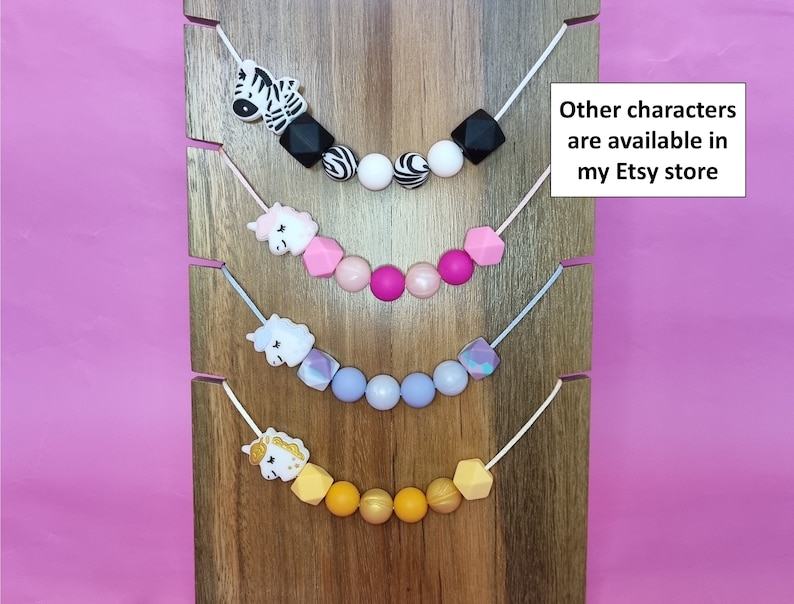Puede incluir: Cuatro collares de dentici&oacute;n de silicona con diferentes formas de animales y geom&eacute;tricas. Los collares se muestran sobre un fondo de madera. El collar superior presenta una cebra, el segundo collar presenta un unicornio, el tercer collar presenta un unicornio y el collar inferior presenta un unicornio. El texto "Other characters are available in my Etsy store" se muestra en el lado derecho de la imagen.
