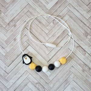 Puede incluir: Un collar de silicona blanco con cuentas negras y amarillas. El collar tiene un dije de ping&uuml;ino.