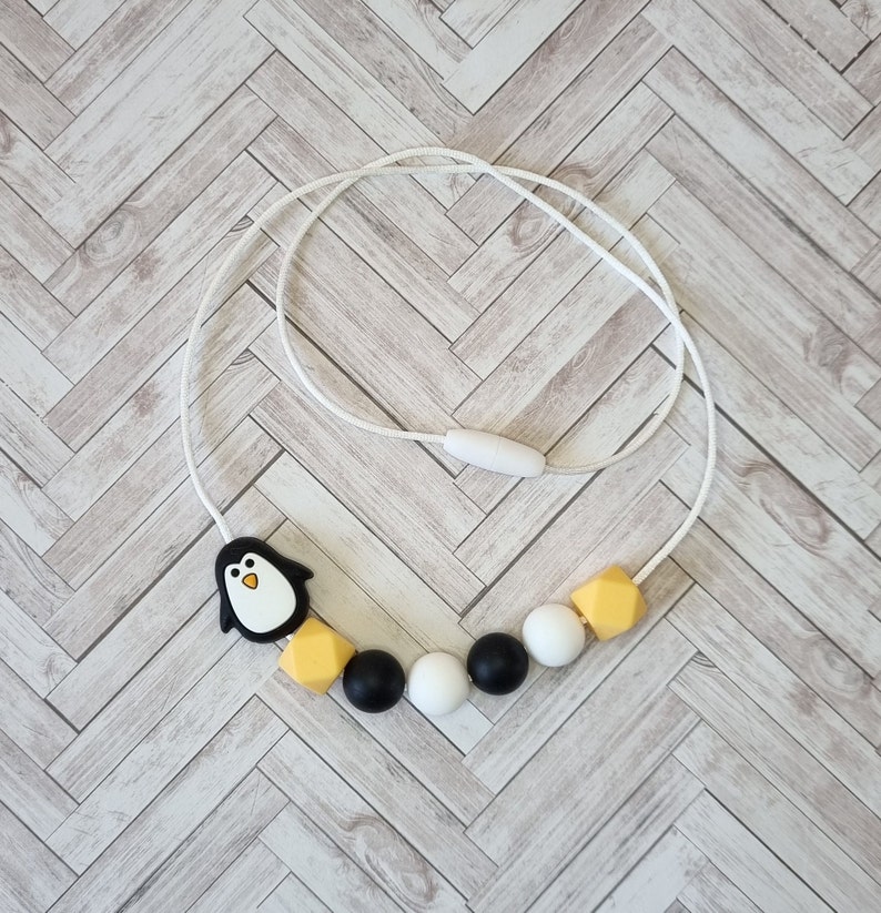 Puede incluir: Un collar de silicona blanco con cuentas negras y amarillas. El collar tiene un dije de ping&uuml;ino.
