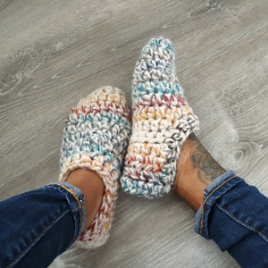 May include: A pair of handmade crochet slippers in cream, teal, red, and yellow. The slippers are thick and cozy, perfect for indoor wear. The yarn has a textured appearance, and the slippers are shown on a light gray wooden floor.