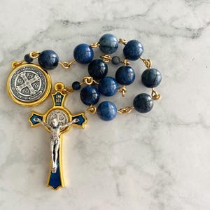 May include: A rosary with blue gemstone beads and gold-colored metal accents. The cross features a silver figure of Jesus and blue enamel detailing. A round medallion with text is also present.