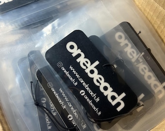May include: A stack of black car air fresheners with the text "onebeach" and the website address "www.onebeach.lt" printed on them. The air fresheners are packaged in clear plastic bags.