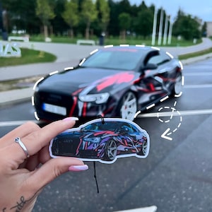 May include: A black and white car air freshener with a colorful abstract design. The air freshener is hanging from a string and is being held by a hand.