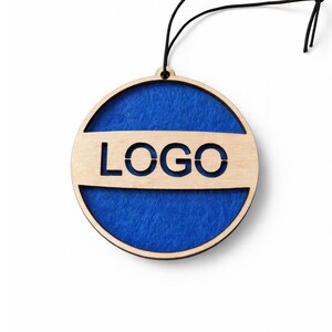 Puede incluir: Adorno redondo de madera con un centro de fieltro azul y la palabra "LOGO" recortada. El adorno tiene una cuerda negra para colgar. La madera tiene un acabado natural.