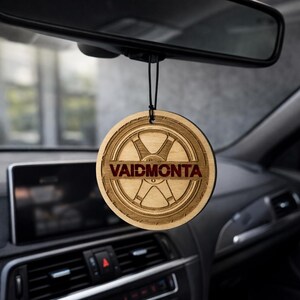 Puede incluir: Un ambientador de coche redondo de madera con la palabra "VAIDMONTA" en letras rojas. El diseño presenta una rueda. Cuelga de una cuerda negra, frente al interior de un coche.