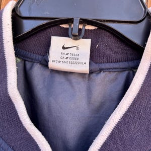 May include: Navy blue jacket with white striped trim on the collar and sleeves. The jacket has a zipper closure and a tag with the Nike logo and product information.