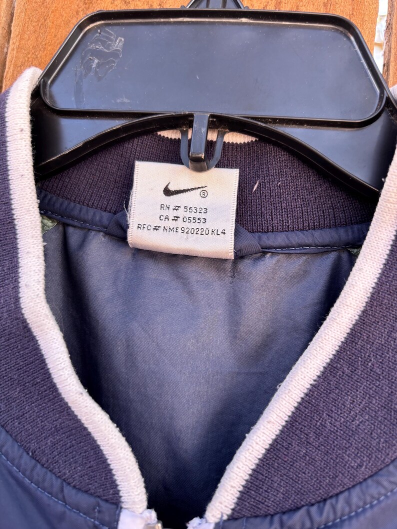 May include: Navy blue jacket with white striped trim on the collar and sleeves. The jacket has a zipper closure and a tag with the Nike logo and product information.
