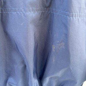 May include: Close-up of a navy blue nylon jacket. The jacket has a light blue stitched seam across the upper back and a white elastic hem. There are several light-colored stains on the jacket.