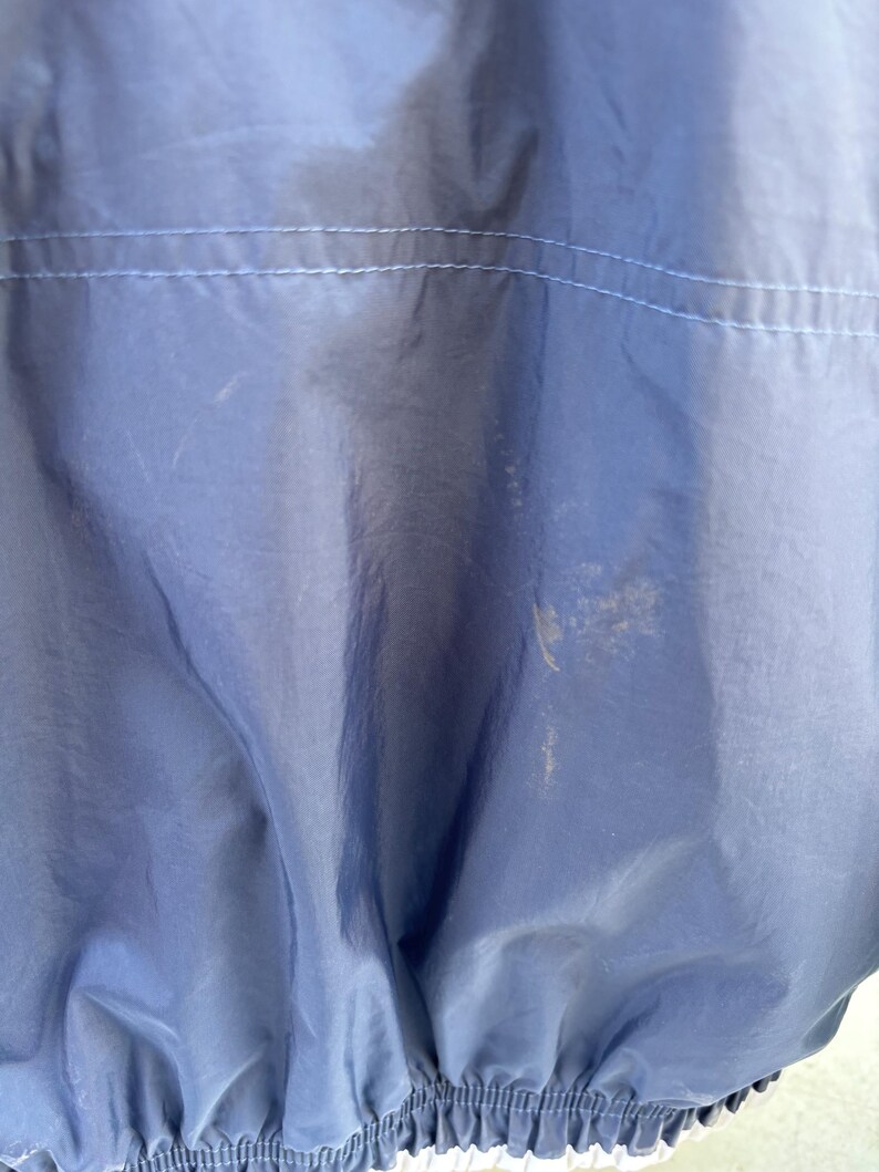 May include: Close-up of a navy blue nylon jacket. The jacket has a light blue stitched seam across the upper back and a white elastic hem. There are several light-colored stains on the jacket.
