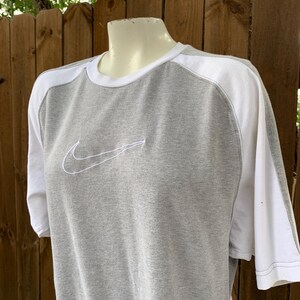 Puede incluir: Camiseta gris y blanca de manga corta con el logotipo de Nike swoosh bordado en blanco en el pecho. La camiseta tiene cuello redondo y paneles blancos en los hombros y las mangas.
