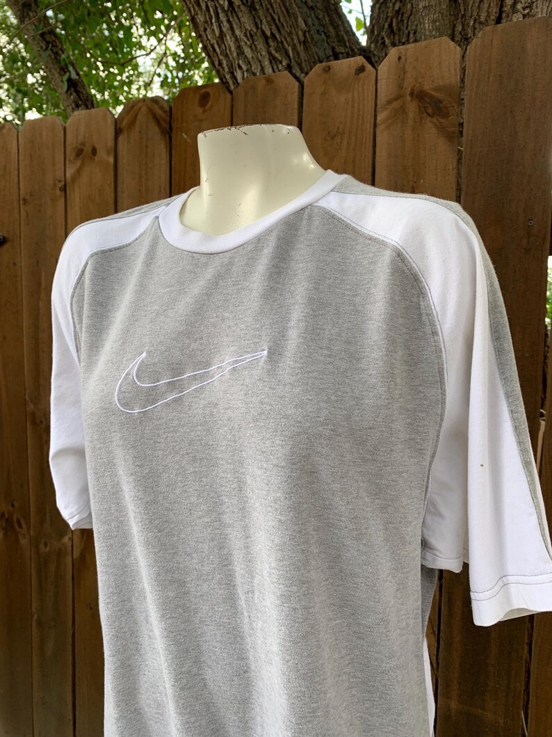 Puede incluir: Camiseta gris y blanca de manga corta con el logotipo de Nike swoosh bordado en blanco en el pecho. La camiseta tiene cuello redondo y paneles blancos en los hombros y las mangas.