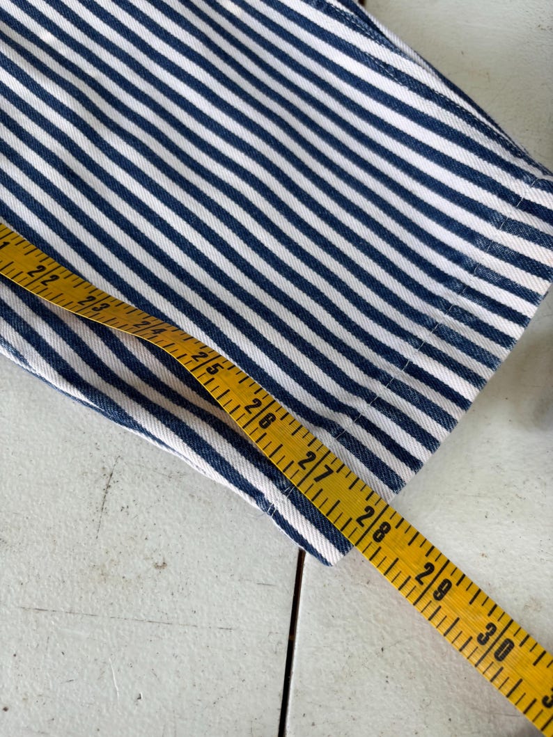 Puede incluir: Primer plano de una tela a rayas azules y blancas. Una cinta m&eacute;trica amarilla se coloca sobre la tela, mostrando medidas en cent&iacute;metros. La tela parece ser mezclilla o un material similar.