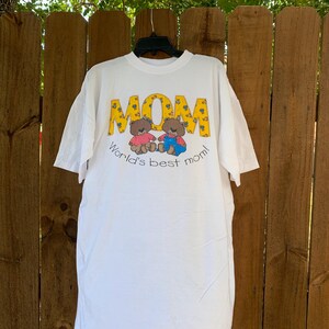以下が含まれることがあります： 2匹のテディベアと「World's best mom!」の文字が書かれた黄色い「MOM」のグラフィックがプリントされた白いTシャツ