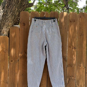 Puede incluir: Pantalones cortos de cintura alta con rayas verticales azul marino y blancas. Los pantalones est&aacute;n colgados de una percha negra contra una valla de madera. Los bolsillos traseros y los botones son visibles.