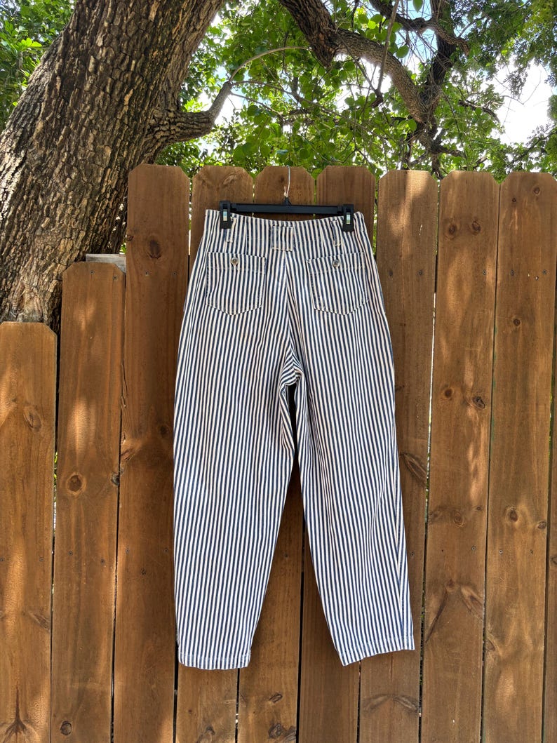 Puede incluir: Pantalones cortos de cintura alta con rayas verticales azul marino y blancas. Los pantalones est&aacute;n colgados de una percha negra contra una valla de madera. Los bolsillos traseros y los botones son visibles.