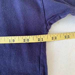 May include: Close-up of a dark blue t-shirt with a faded purple ombre effect. A measuring tape is placed along the bottom edge of the shirt, showing measurements from 19 to 23 inches.
