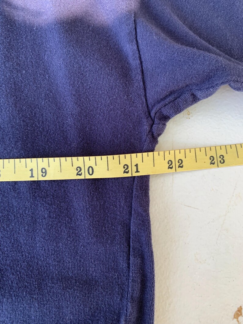 May include: Close-up of a dark blue t-shirt with a faded purple ombre effect. A measuring tape is placed along the bottom edge of the shirt, showing measurements from 19 to 23 inches.