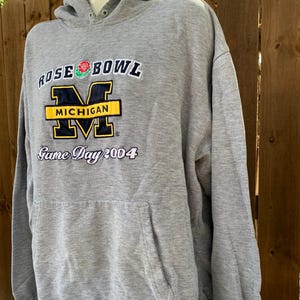 May include: A gray hooded sweatshirt with a front pocket and the text "ROSE BOWL MICHIGAN Game Day 2004". The text is in white, black, yellow, and blue. The sweatshirt is displayed on a mannequin.