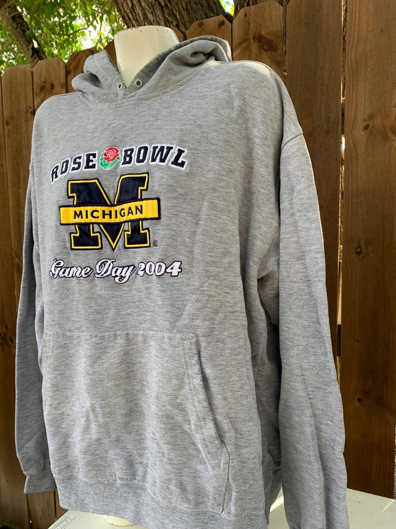 May include: A gray hooded sweatshirt with a front pocket and the text "ROSE BOWL MICHIGAN Game Day 2004". The text is in white, black, yellow, and blue. The sweatshirt is displayed on a mannequin.