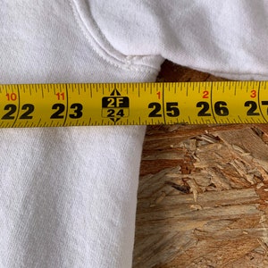 May include: Close-up of a white cotton sweatshirt with a yellow measuring tape showing 22 to 27 inches.
