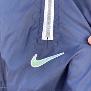 May include: Navy blue windbreaker jacket with a white and gray zipper and a light green Nike logo. The jacket has a simple design with a classic athletic aesthetic.
