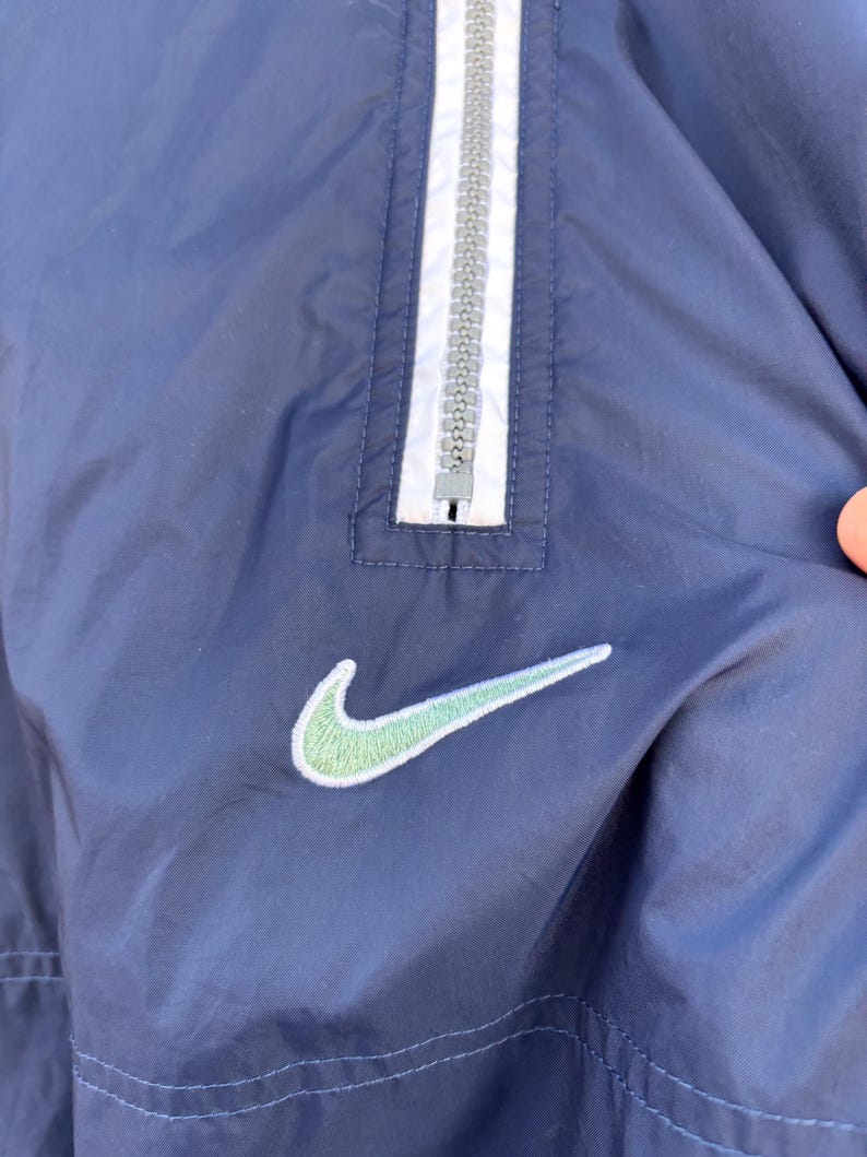May include: Navy blue windbreaker jacket with a white and gray zipper and a light green Nike logo. The jacket has a simple design with a classic athletic aesthetic.