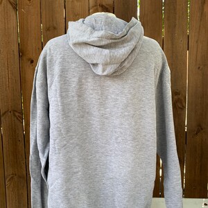 May include: A heather gray hoodie with a drawstring hood and long sleeves. The back view of the sweatshirt is shown against a wooden fence and a tree.