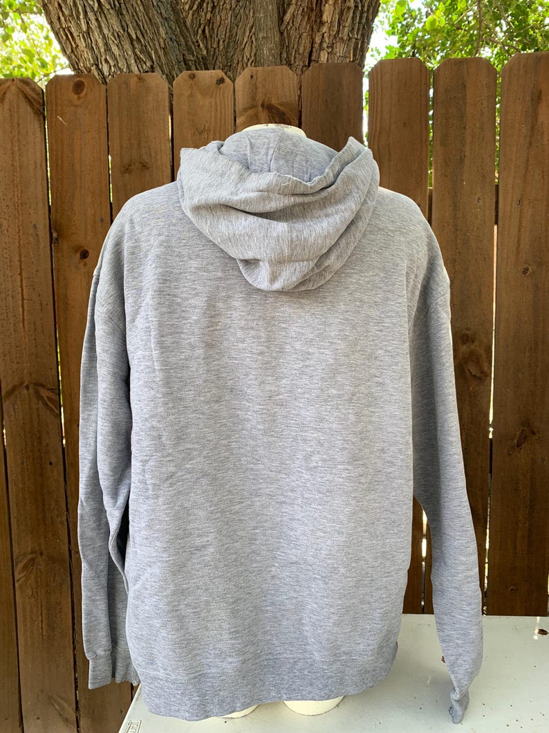 May include: A heather gray hoodie with a drawstring hood and long sleeves. The back view of the sweatshirt is shown against a wooden fence and a tree.