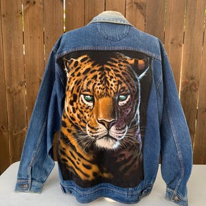 May include: A denim jacket with a large, detailed graphic of a leopard's face on the back. The leopard has green eyes and a spotted coat. The jacket is a medium wash blue with button closures and pockets.