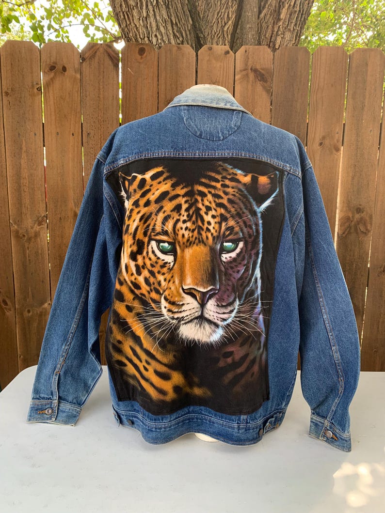 May include: A denim jacket with a large, detailed graphic of a leopard's face on the back. The leopard has green eyes and a spotted coat. The jacket is a medium wash blue with button closures and pockets.