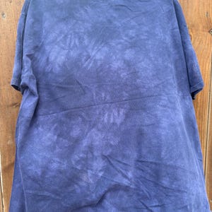 May include: A navy blue tie-dye t-shirt hanging on a black hanger. The shirt has a crew neck and short sleeves. The tie-dye pattern is a mix of dark and light blue hues, creating a textured effect.