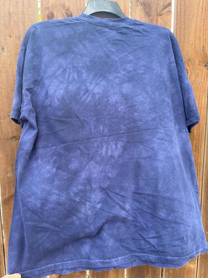 May include: A navy blue tie-dye t-shirt hanging on a black hanger. The shirt has a crew neck and short sleeves. The tie-dye pattern is a mix of dark and light blue hues, creating a textured effect.