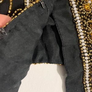 May include: Close-up of a black garment with gold and white beading. The garment is lined with black fabric and has a hook and eye closure.