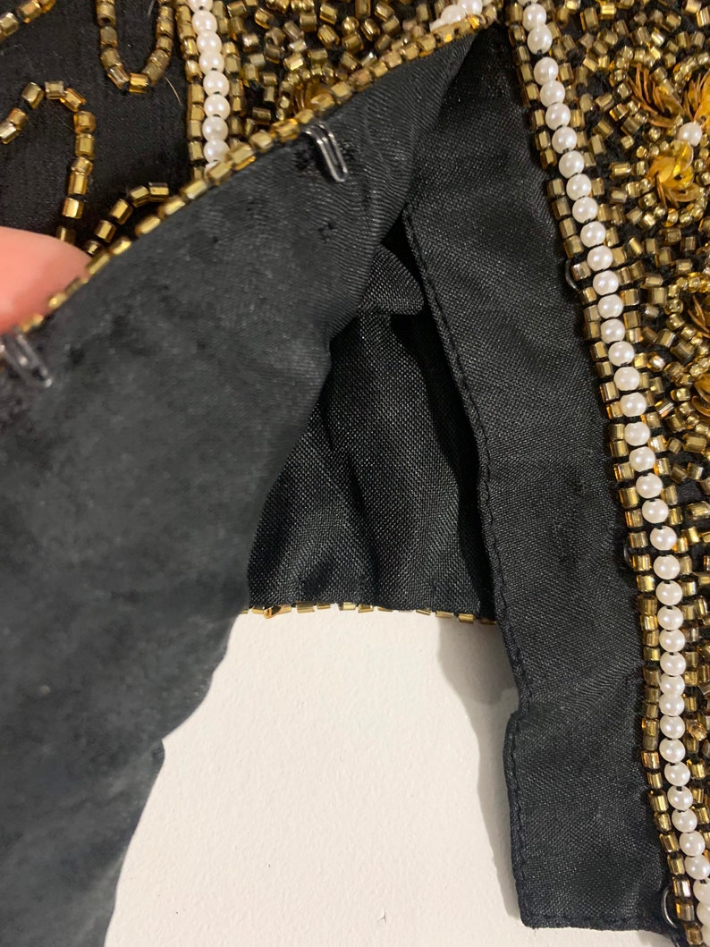 May include: Close-up of a black garment with gold and white beading. The garment is lined with black fabric and has a hook and eye closure.