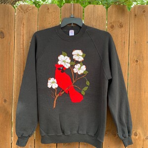 Op de afbeelding: Zwart sweatshirt met een rode kardinaalvogel op een tak met witte bloemen. Het vogel- en bloemendesign is geborduurd op de voorkant van de top met lange mouwen. Het sweatshirt hangt aan een zwarte hanger.
