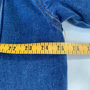 May include: Close-up of blue denim fabric with visible stitching. A yellow measuring tape shows a length of approximately 22 to 26 inches. The denim has a classic, textured appearance.
