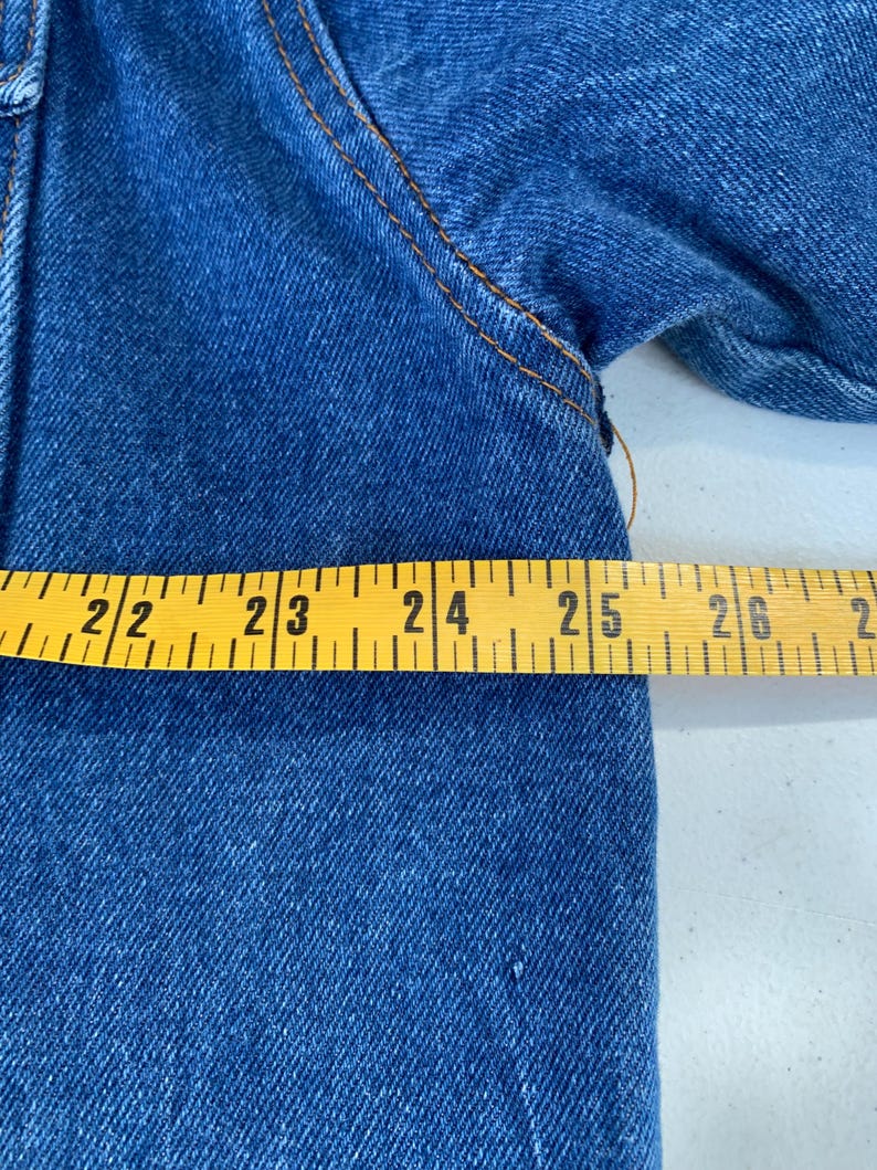 May include: Close-up of blue denim fabric with visible stitching. A yellow measuring tape shows a length of approximately 22 to 26 inches. The denim has a classic, textured appearance.