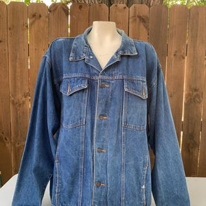 May include: A medium-wash denim jacket with a classic design. The jacket features a collared neckline, button-down closure, two chest pockets, and two side pockets. The jacket is displayed on a mannequin against a wooden fence background.