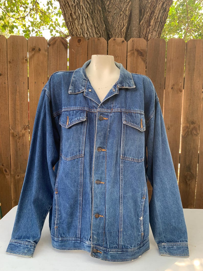 May include: A medium-wash denim jacket with a classic design. The jacket features a collared neckline, button-down closure, two chest pockets, and two side pockets. The jacket is displayed on a mannequin against a wooden fence background.