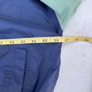 May include: A navy blue and light green jacket with a white trim. The jacket is being measured with a yellow measuring tape. The jacket has a pocket on the front. The jacket is made of a lightweight material.